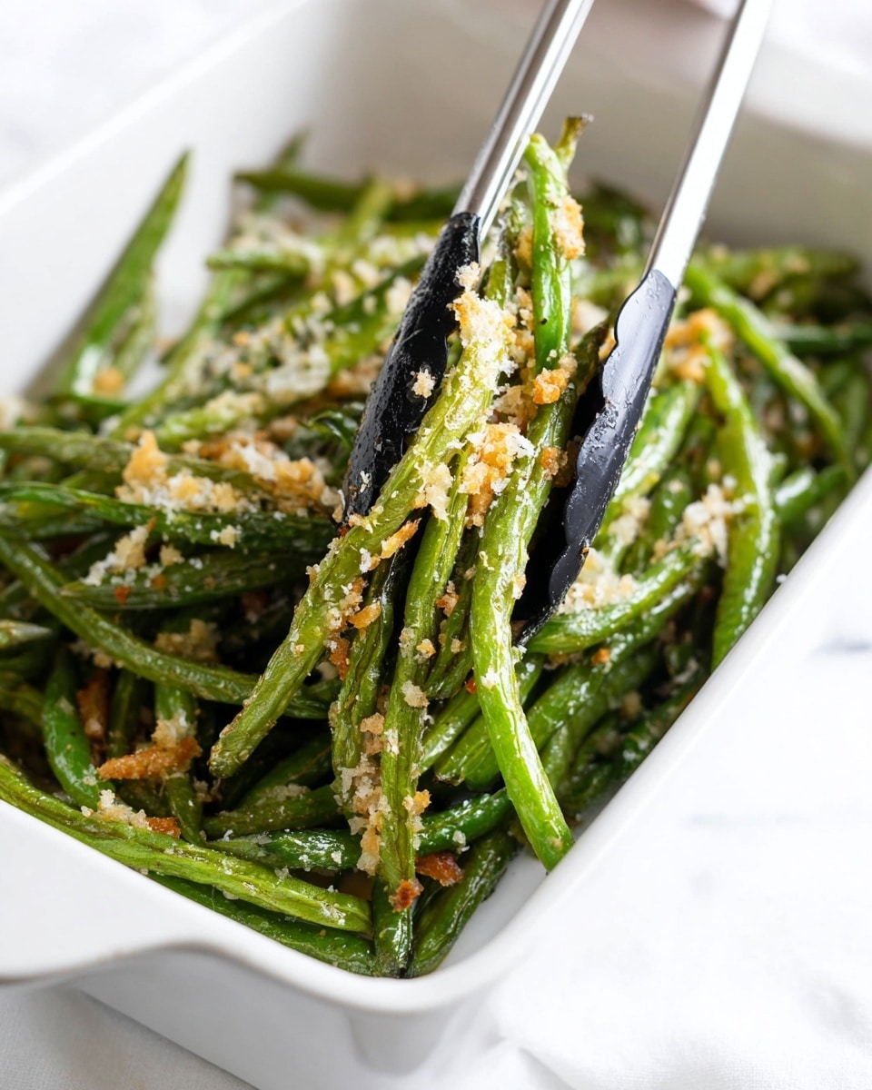 The image shows a white rectangular dish filled with roasted green beans that have a slightly shiny, cooked texture and patches of light browning. The green beans are topped with small breadcrumbs or grated cheese pieces scattered all over. A pair of black tongs with silver handles is picking up a bunch of beans from the dish. The background surface is white with a marble texture. photo taken with an iphone --ar 4:5 --v 7