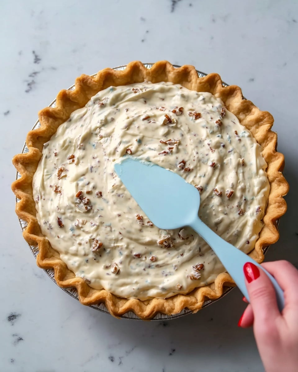 A round pie with a golden brown crust that has a crimped edge, filled with a creamy white mixture containing small brown bits evenly spread throughout. A woman's hand with red nail polish holds a light blue spatula gently spreading the filling across the top. The pie sits on a white marbled surface. photo taken with an iphone --ar 4:5 --v 7