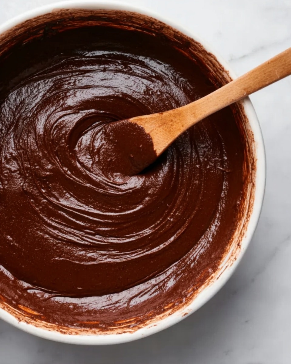 The image shows a close-up of a white bowl filled with thick, smooth chocolate batter. The batter is dark brown and shiny with a slightly textured surface, swirled softly in the bowl. A wooden spoon with traces of the batter rests partially submerged in the mixture, positioned toward the top right side. The bowl sits on a white marbled surface. photo taken with an iphone --ar 4:5 --v 7