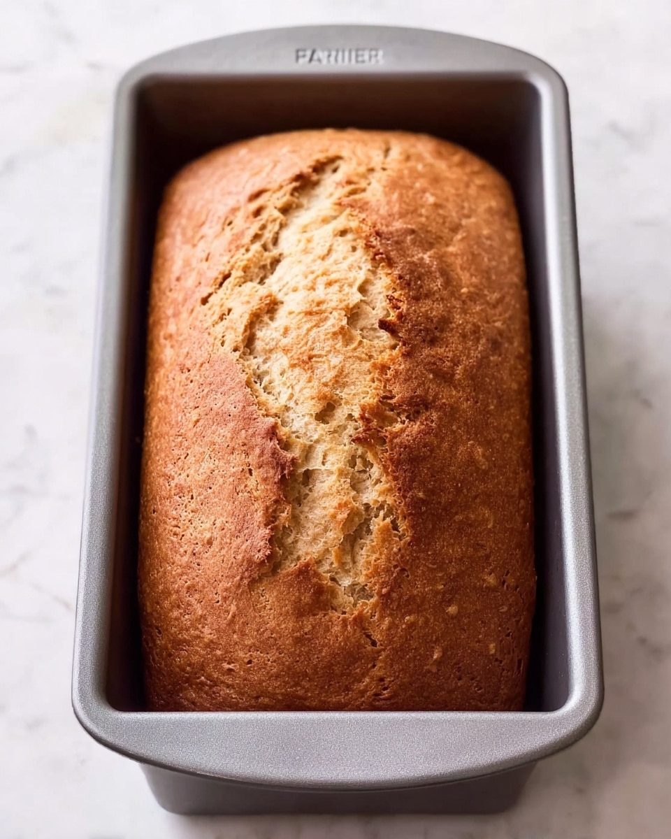 A golden brown loaf of bread with a cracked top surface lies inside a grey metal baking pan. The loaf’s texture shows a slightly rough, crisp crust with some darker brown areas, indicating a well-baked finish. The pan is rectangular with smooth edges, resting on a white marbled surface that adds a clean and bright background to the image. photo taken with an iphone --ar 4:5 --v 7