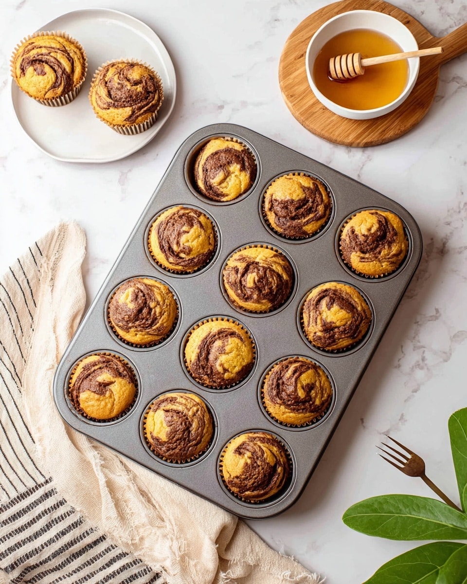 A metal muffin tray holds ten golden brown muffins with dark brown swirls on top, placed on a white marbled surface. To the top left, two muffins sit on a white plate and wooden board. On the right, there is a white bowl filled with honey and a wooden honey dipper resting inside. A beige cloth with black stripes is spread out on the left side under the muffin tray. At the bottom right corner, a green leafy plant and a wooden fork are visible. Photo taken with an iphone --ar 4:5 --v 7
