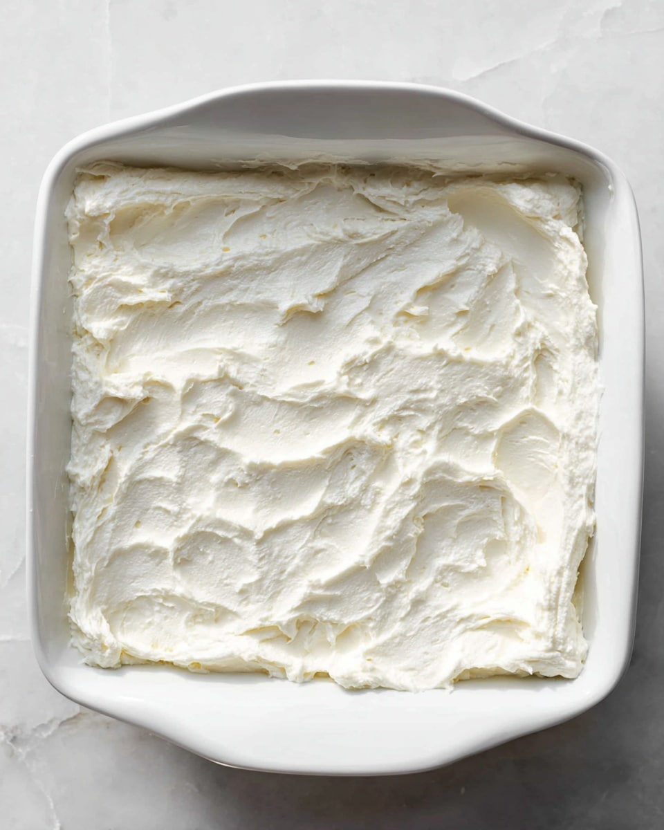 A square white ceramic dish filled with a thick, fluffy white layer of cream spread evenly across the surface, showing some soft, smooth swirls and slight peaks. The cream looks light and textured, covering the entire inside of the dish, which sits on a white marbled surface. The edges of the dish have soft curves and the cream layer appears fresh and airy. photo taken with an iphone --ar 4:5 --v 7