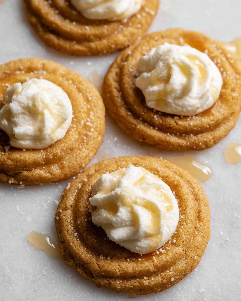 The image shows four round cookies in a close diagonal line. Each cookie is light brown with a slightly crinkled texture and soft, swirled ridges. On top of each cookie, there is a dollop of white cream or frosting, slightly uneven and soft in appearance. The cookies rest on a white marbled surface with some tiny scattered crumbs and drops of syrup around them. The overall look is warm and inviting with a soft focus on the front cookie. photo taken with an iphone --ar 4:5 --v 7