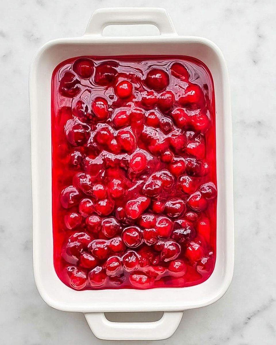 The image shows a white rectangular dish on a white marbled surface. Inside the dish is a single thick layer of bright, glossy red cherry sauce filled with whole cherries. The sauce has a smooth, shiny texture with some swirls and slight ripples, making the cherries stand out with their round shapes and deeper red color against the lighter red sauce. The dish handle is visible at the top and bottom edges. Photo taken with an iphone --ar 4:5 --v 7