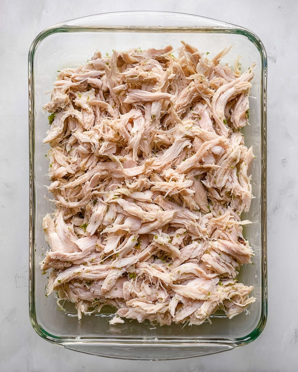 Chicken Cobbler Recipe 4 A clear glass baking dish holds one layer of shredded pale pink chicken scattered evenly across the bottom. A slight shine on the chicken shows moisture, while bits of green seasoning peek through underneath. The dish sits on a surface with a clean white marbled texture. The chicken strands are uneven and loose, giving a soft texture look. Photo taken with an iphone --ar 4:5 --v 7