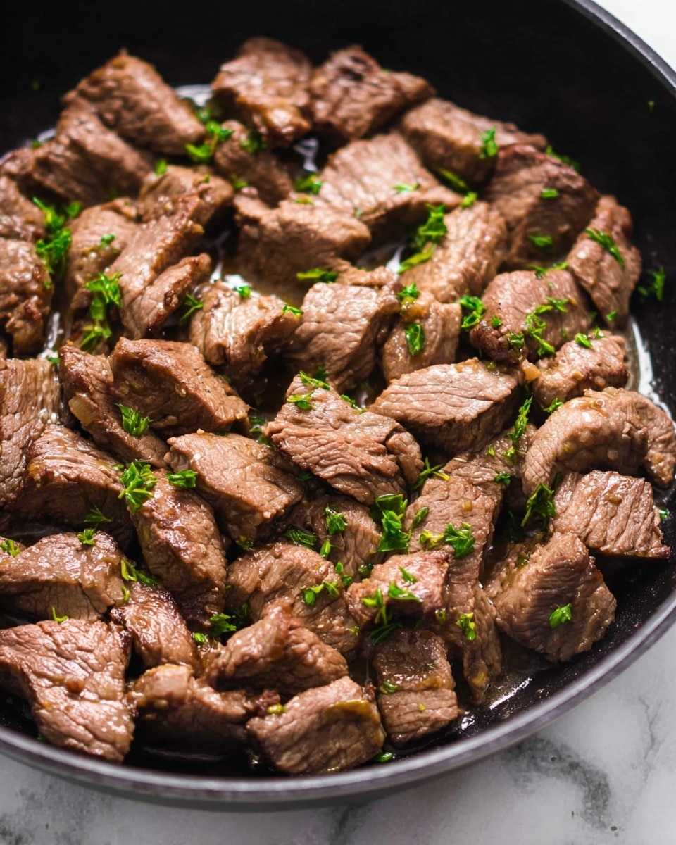 This image shows many small chunks of cooked beef in a black pan. The beef pieces are brown with a soft texture and slightly uneven shapes. They are scattered closely together, and some small green herb pieces are sprinkled on top for color. The beef appears moist with a bit of shiny sauce or liquid around it, giving it a juicy look. The pan rests on a white marbled surface. photo taken with an iphone --ar 4:5 --v 7