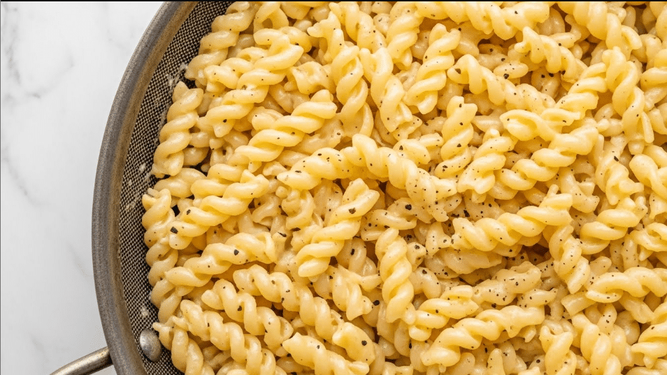 This image shows a close-up of a large pan filled with cooked spiral pasta coated in creamy, pale yellow cheese sauce with a smooth and slightly thick texture. The sauce evenly covers every twist of the pasta, giving it a shiny, soft look. Small black specks of seasoning are scattered throughout the sauce. The pan has a metal rim and mesh sides, hinting at it being used for draining. The background surface is a white marbled texture. Photo taken with an iphone --ar 4:5 --v 7