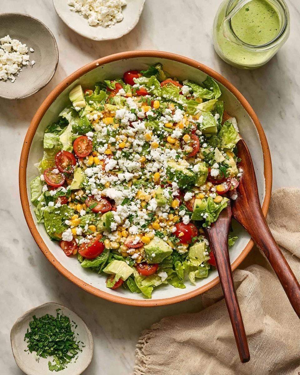 A large white bowl with a brown rim holds a fresh salad made of green chopped lettuce as the base layer, mixed with yellow corn kernels, halved red cherry tomatoes, and diced green avocado pieces. Crumbled white cheese is scattered evenly on top, adding a soft texture and bright contrast. Two wooden salad spoons with dark handles rest inside the bowl, partially lifting the salad. Around the bowl, there is a small white bowl with chopped green herbs, another with crumbled white cheese, and a clear glass jar filled with a light green dressing. All items are placed on a white marbled surface with a beige cloth nearby. photo taken with an iphone --ar 4:5 --v 7