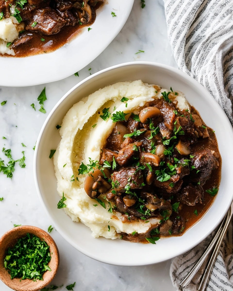 The image shows a close-up of a white bowl filled with two layers: the bottom layer is smooth, white mashed potatoes with a creamy texture and slight swirls around the edge, topped with a rich, dark brown beef stew that has chunks of tender meat, small round onions, and cooked mushrooms visible within the thick sauce. The stew is garnished with fresh green parsley leaves scattered evenly on top, adding a pop of color. The bowl sits on a white marbled surface sprinkled with a few parsley leaves. Nearby, there is a small wooden bowl filled with chopped parsley and part of a white plate with more mashed potatoes and stew visible, along with a silver fork and a striped cloth napkin. photo taken with an iphone --ar 4:5 --v 7