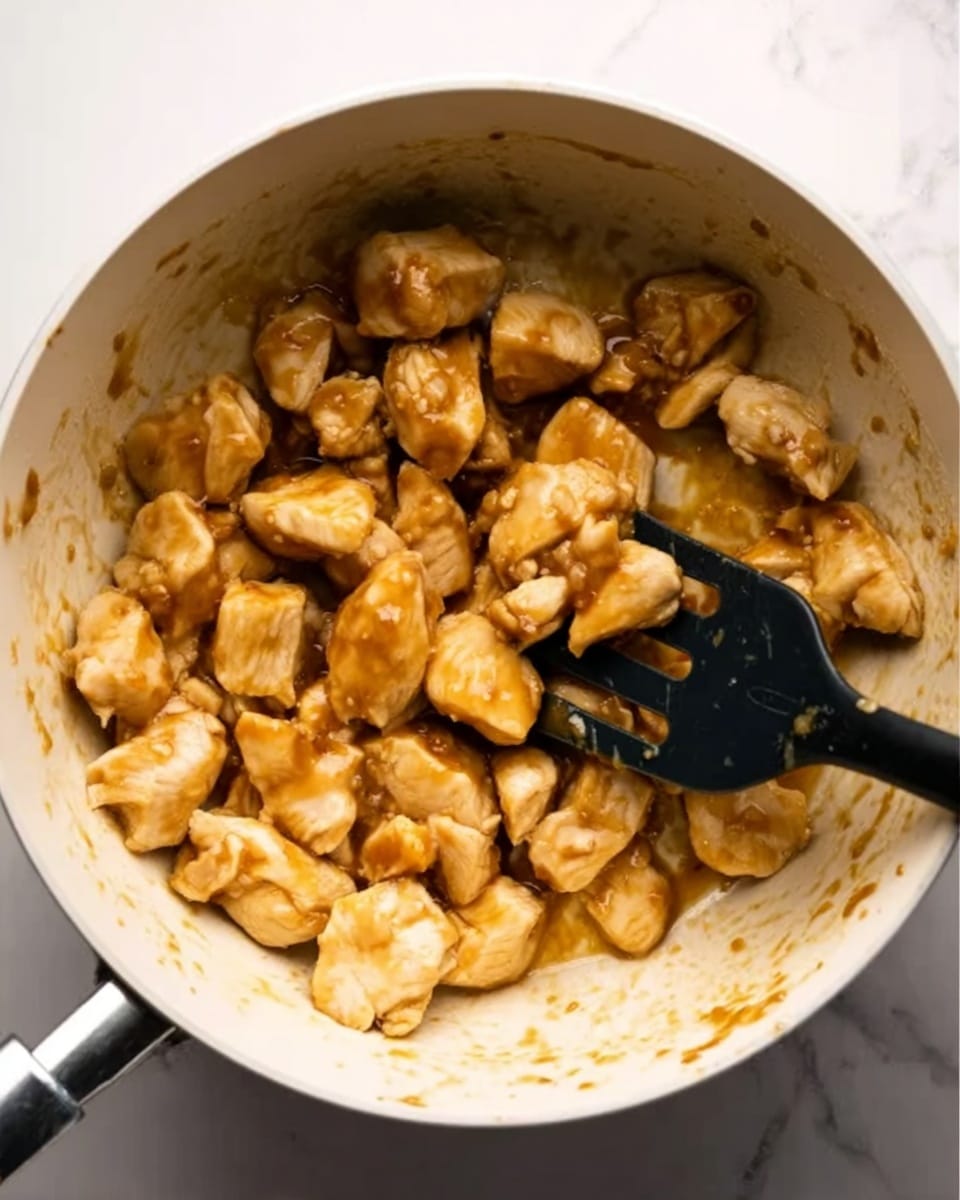 A white pot filled with small pieces of cooked chicken coated in a light brown sauce. The chicken pieces are chunky and scattered mostly towards the bottom half of the pot. A black slotted spatula is resting inside the pot on the right side, touching some chicken pieces. The pot is placed on a white marbled surface. Photo taken with an iphone --ar 4:5 --v 7