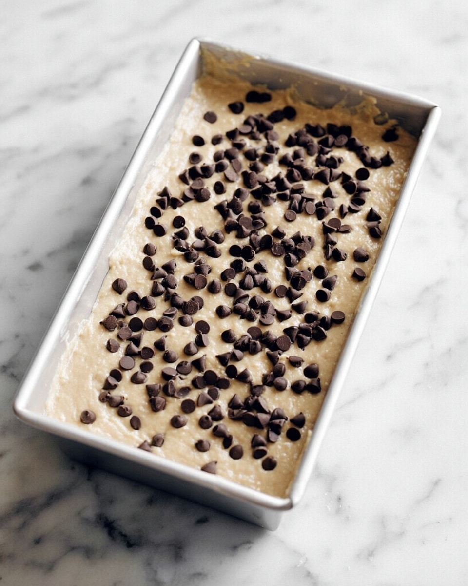 A rectangular white baking pan filled with light beige batter that looks smooth and thick, topped evenly with many small dark brown chocolate chips scattered all over the surface, placed on a white marbled textured surface photo taken with an iphone --ar 4:5 --v 7
