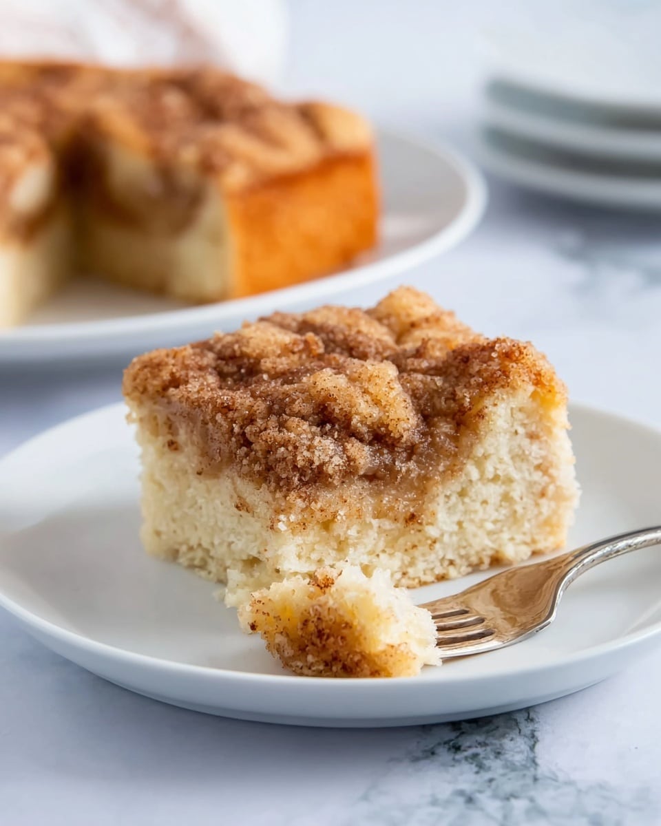 A piece of cinnamon coffee cake on a white plate shows two layers: the bottom layer is light beige, soft and moist cake, while the top layer is a golden brown crumbly cinnamon sugar topping with a slightly crispy texture. A small square piece of cake is on a silver fork resting on the plate, showing the soft inside and crumbly top. In the background, a second piece of the cake is on another white plate, and the surface has a white marbled texture. The photo taken with an iphone --ar 4:5 --v 7