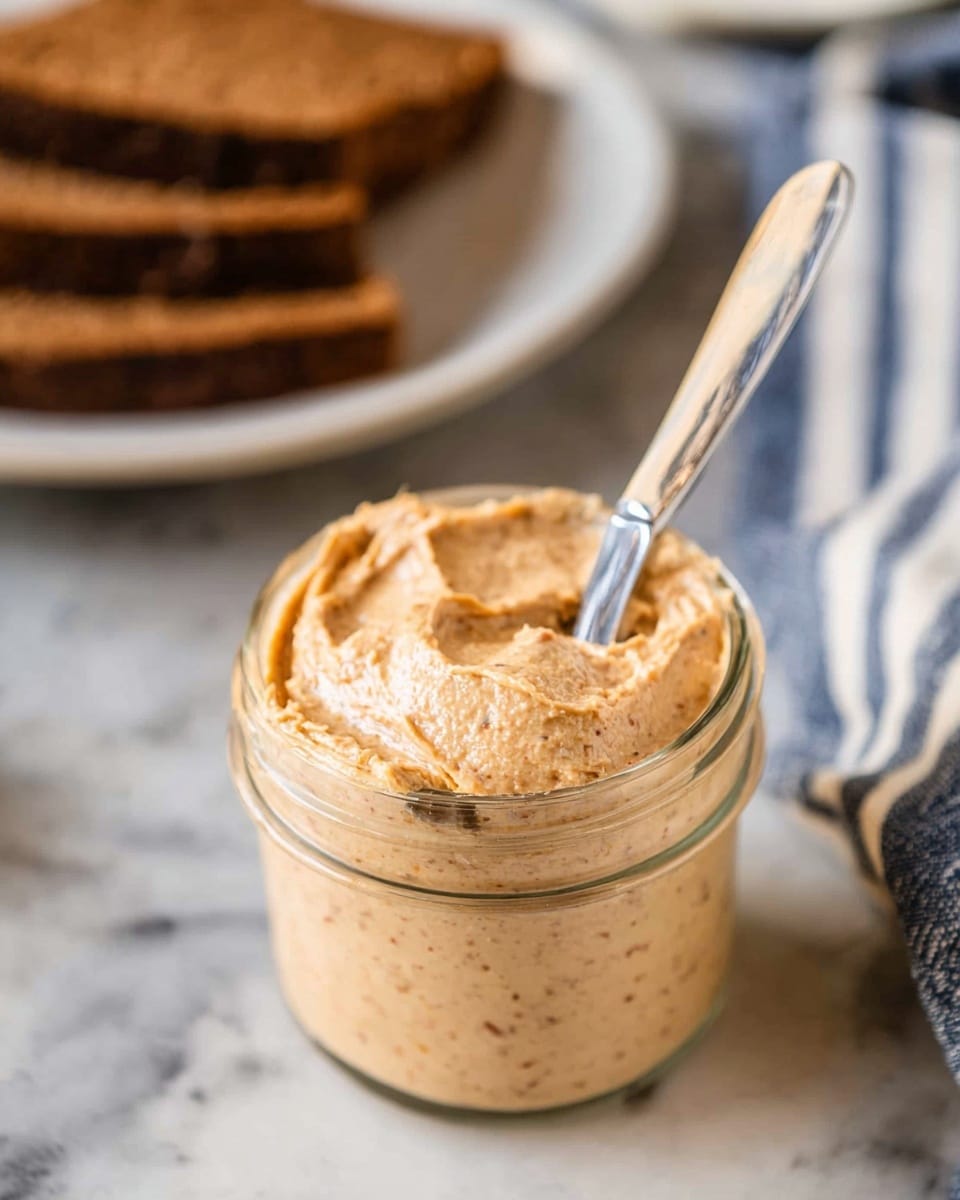 A clear glass jar filled with a creamy, light brown spread that has a smooth texture with tiny darker specks mixed in, with a silver butter knife resting on top of the spread inside the jar; in the background, there is a blurred white plate containing thick slices of dark brown bread, all placed on a white marbled surface with a blue and white striped cloth nearby. photo taken with an iphone --ar 4:5 --v 7
