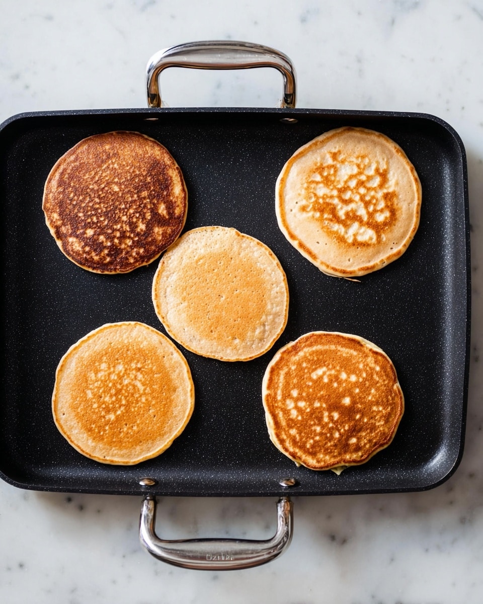 Five small round pancakes are cooking on a black pan with silver handles on each side, placed on a white marbled surface. The pancakes vary in color and texture: one pancake near the bottom left is darker brown with a crispy look, two pancakes in the bottom middle and bottom right are lighter with spots and a slightly bubbly texture, the pancake at the top left is light brown and smooth, and the pancake at the top right has a more even golden-brown ring and a patterned surface. The pan's surface is smooth and non-stick. photo taken with an iphone --ar 4:5 --v 7