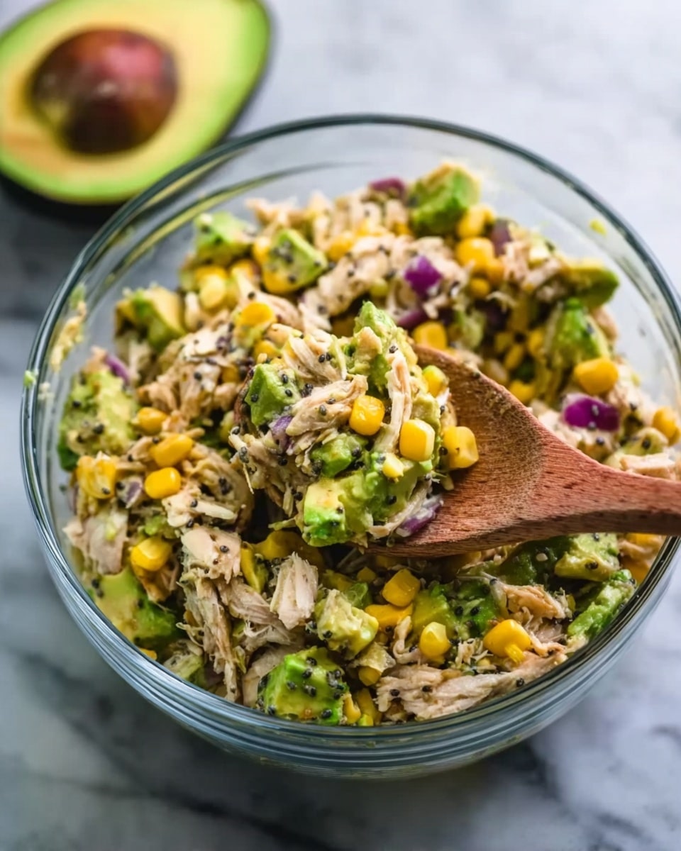 A clear glass bowl filled with a colorful salad sits on a white marbled surface. The salad has several layers: at the bottom, pieces of light brown shredded chicken; mixed throughout are bright yellow corn kernels, chopped green avocado slices, small purple onion bits, and black seeds that look like chia or poppy seeds. A woman's hand is holding a wooden spoon on the right side, lifting some of the salad. In the background and slightly blurred, a halved avocado with a green outer skin and yellow flesh is visible. The photo taken with an iphone --ar 4:5 --v 7