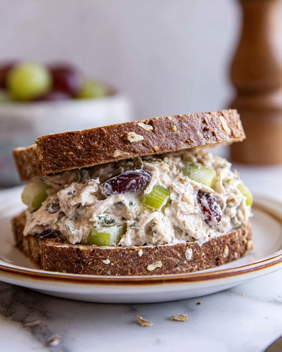 A thick sandwich with two slices of dark brown multigrain bread filled with a creamy mixture that includes white shredded chicken, light green celery pieces, and dark red grapes, all mixed with a white creamy dressing. The sandwich sits on a white plate with a thin brown rim, placed on a white marbled surface. The background is softly blurred, hinting at a bowl and a wooden pepper grinder. The sandwich looks fresh and hearty with a rough textured crust full of oats and seeds on the bread. Photo taken with an iphone --ar 4:5 --v 7