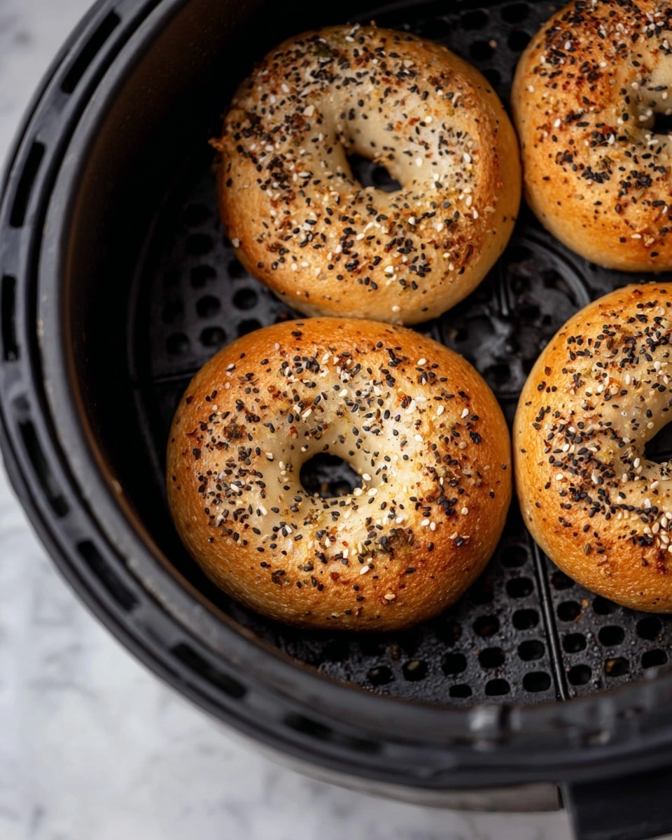 Three round bagels sit inside a black air fryer basket with a grid pattern. The bagels are golden brown with a slightly shiny crust, topped with a mix of black sesame seeds, white sesame seeds, and small bits of light brown seasoning, giving a speckled look. The bagels have a firm texture with small cracks visible on their surface. The background shows part of the air fryer rim in focus, and the surface below is a white marbled texture. Photo taken with an iphone --ar 4:5 --v 7