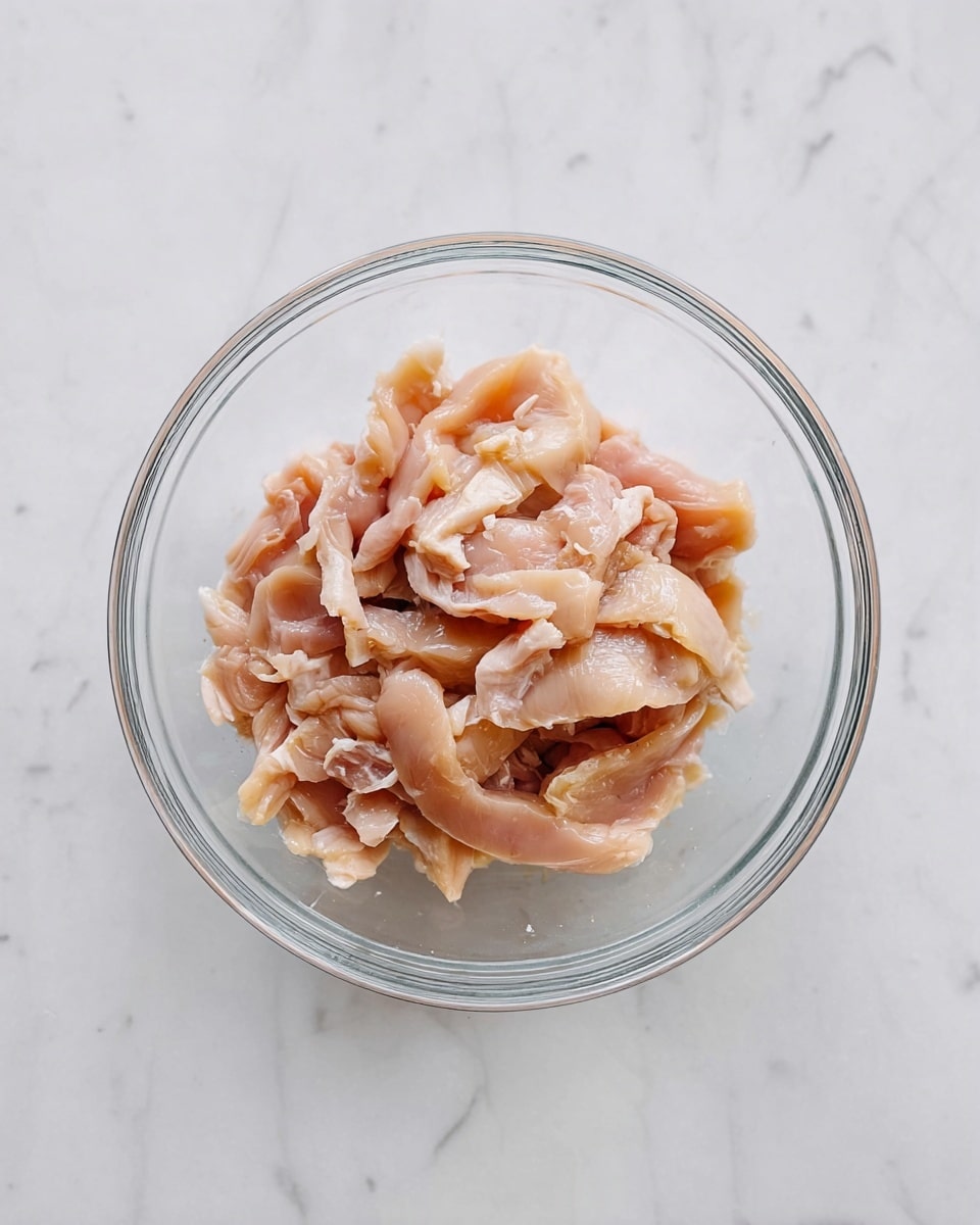 A clear glass bowl sits centered on a white marbled surface. Inside the bowl is one layer of raw chicken strips that are pinkish beige with some white fat bits, showing a soft and slightly shiny texture. The chicken pieces are arranged loosely, filling the bowl with some space around. The overall look is clean and simple, focusing on the fresh raw chicken. Photo taken with an iphone --ar 4:5 --v 7