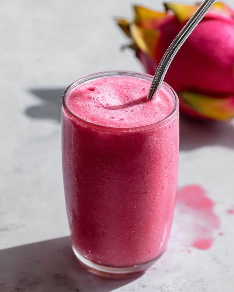 A close-up image of a tall clear glass filled with a bright pink smoothie, showing a smooth and slightly frothy texture with tiny bubbles on the surface. A silver metal straw is placed inside the glass, angled toward the top right. The glass sits on a white marbled surface, with a blurred pink fruit with yellow and green leaves in the background on the right side. There is also a small splash of pink smoothie on the surface near the glass. The lighting highlights the vibrant color and cool condensation on the glass. photo taken with an iphone --ar 4:5 --v 7