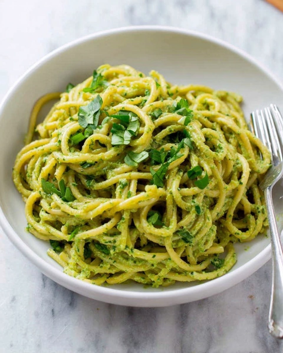 A white bowl filled with thick, yellowish-green noodles mixed evenly with a creamy green sauce, topped with small green herb leaves scattered on top. The bowl sits on a white marbled surface with a silver fork placed near the bowl's edge. The noodles have a slightly smooth and soft texture with visible bits of herbs inside the sauce, making it look fresh and flavorful. Photo taken with an iphone --ar 4:5 --v 7