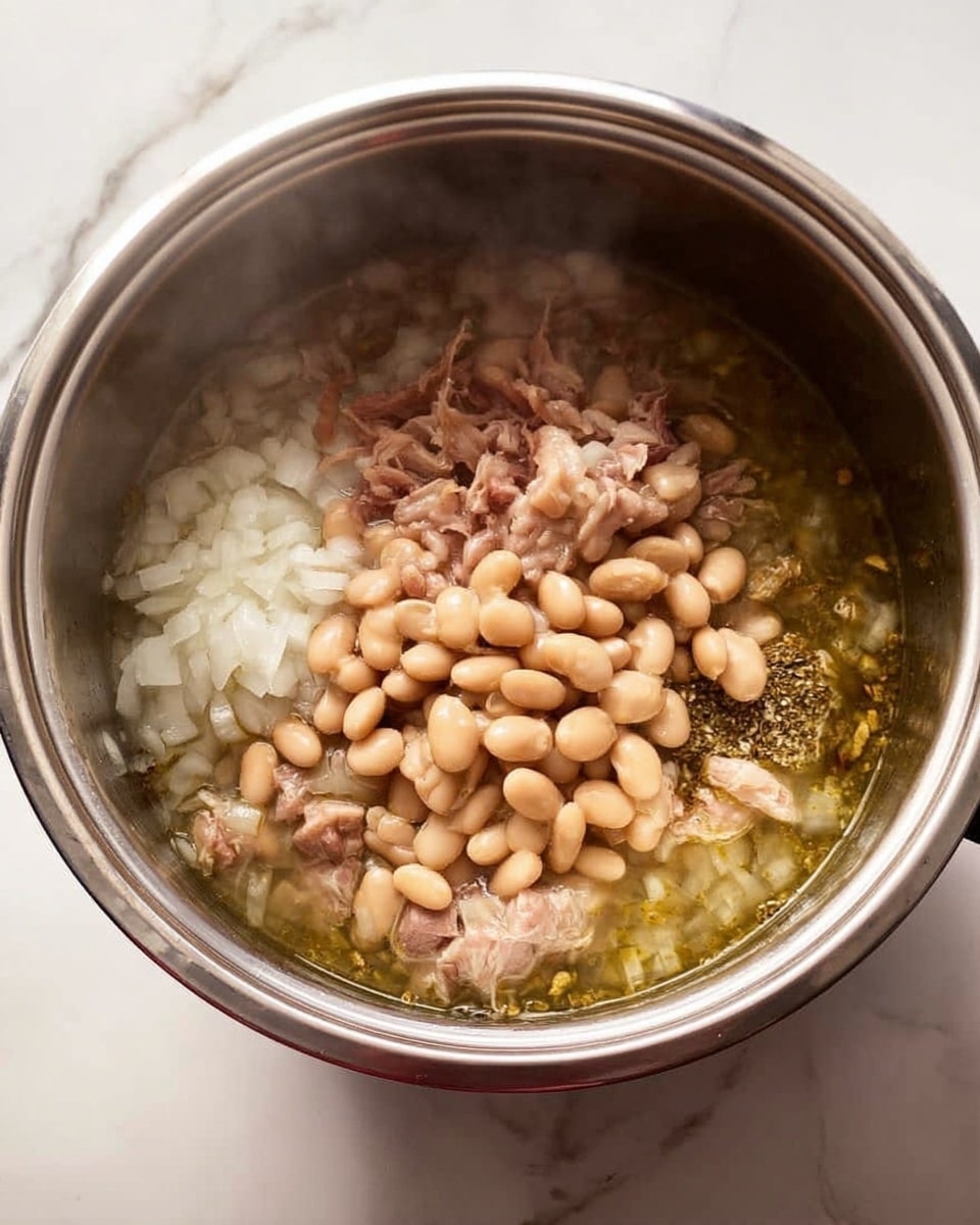 Inside a silver pot, there is a steaming mix of four layers. The bottom layer is a light green liquid broth. Above the broth, finely chopped white onions spread out evenly. On top of the onions, there are small pieces of light brown meat. The top layer has many light tan beans covering the meat. The pot sits on a white marbled surface. photo taken with an iphone --ar 4:5 --v 7