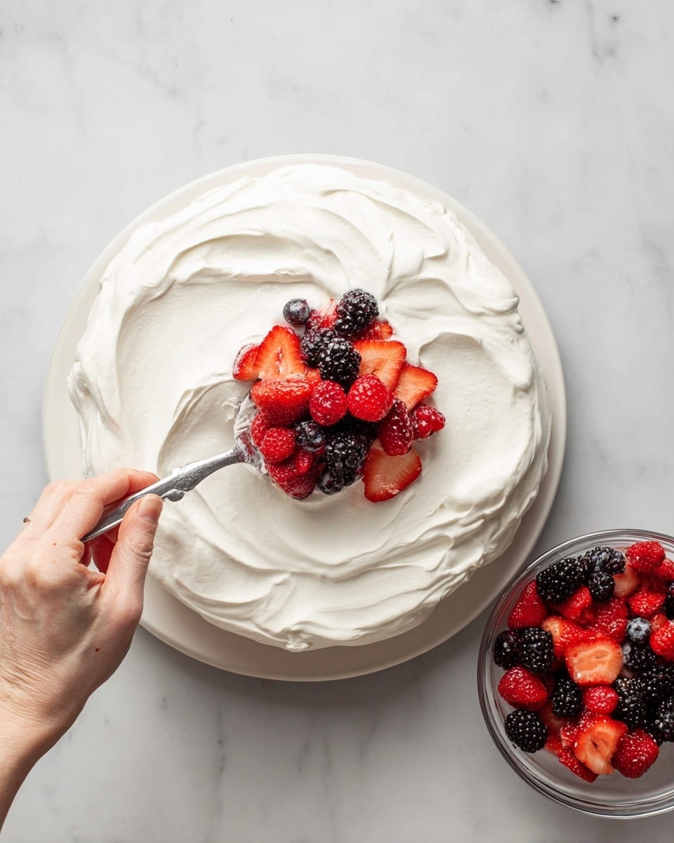 A round dessert sits on a white marbled surface, covered with a thick, smooth layer of white whipped cream that has soft swirls on top. On the right side, a clear glass bowl is filled with mixed berries including bright red strawberries, raspberries, and dark blackberries, all shiny with juice. A woman's hand holds a silver spoon, placing some of the mixed berries on the center of the whipped cream layer. Photo taken with an iphone --ar 4:5 --v 7