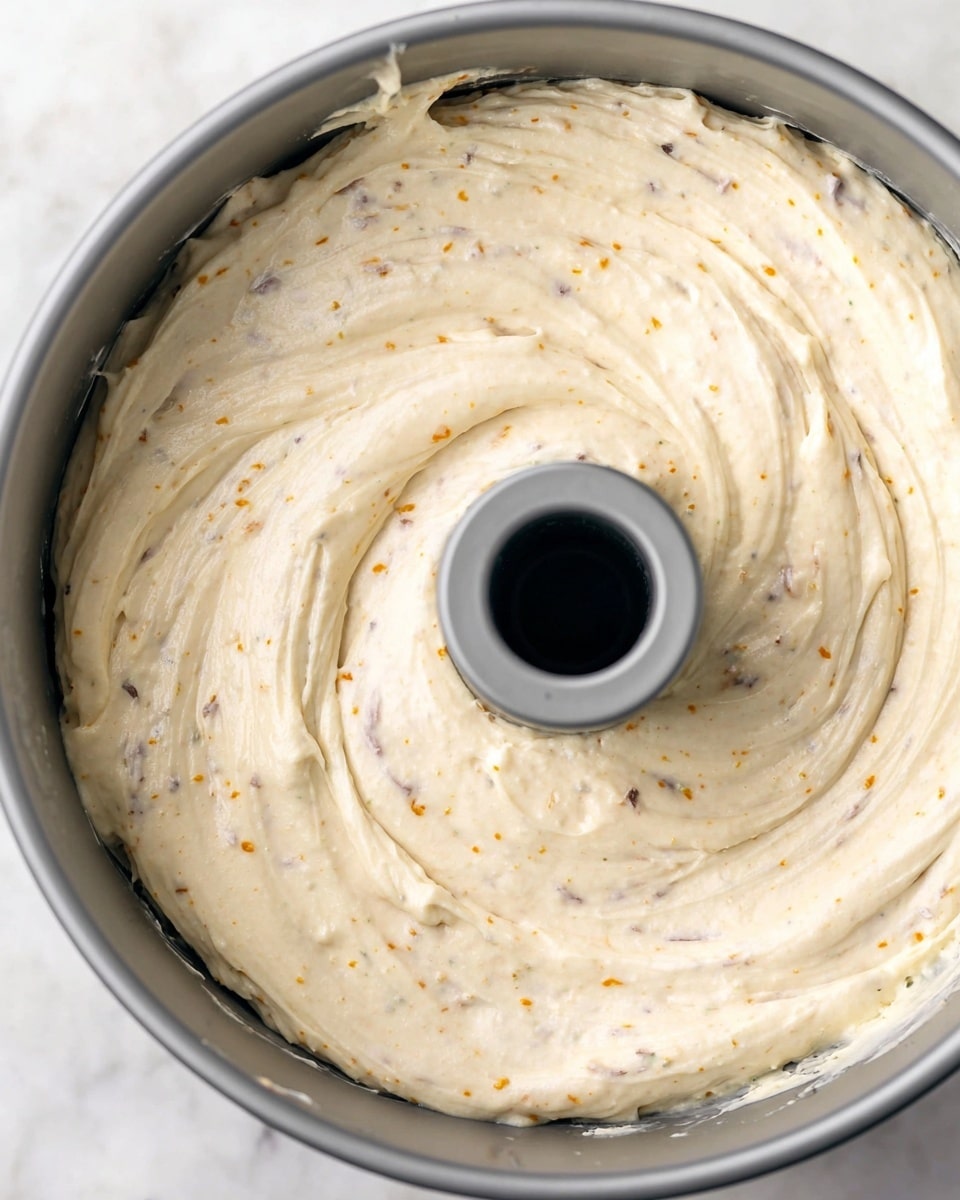 A close-up view of a raw cake batter spread evenly inside a gray bundt pan with a center tube. The batter is pale cream with small dark and orange specks scattered throughout, showing a smooth and thick texture with slight swirls on the surface. The pan sits on a white marbled surface, and the batter fills the pan almost to the edge but does not overflow. Photo taken with an iphone --ar 4:5 --v 7