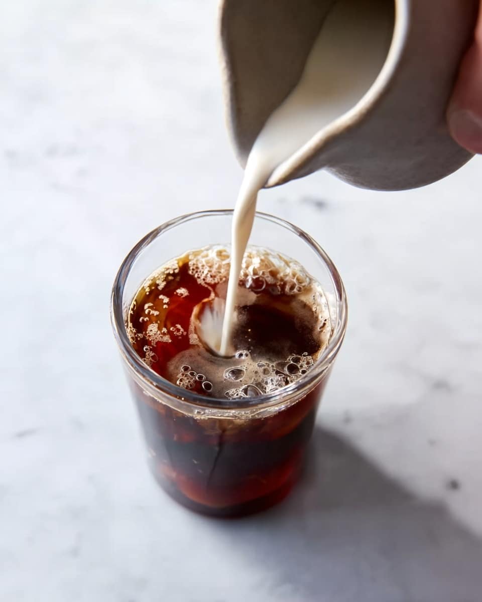 A clear glass filled with dark brown coffee is placed on a white marbled surface. A light gray ceramic pitcher held by a woman’s hand is pouring white cream into the coffee, creating a swirling pattern where the cream mixes with the dark liquid. The top view shows bubbles forming on the coffee surface and the contrast between the dark coffee and the creamy swirl. Photo taken with an iphone --ar 4:5 --v 7