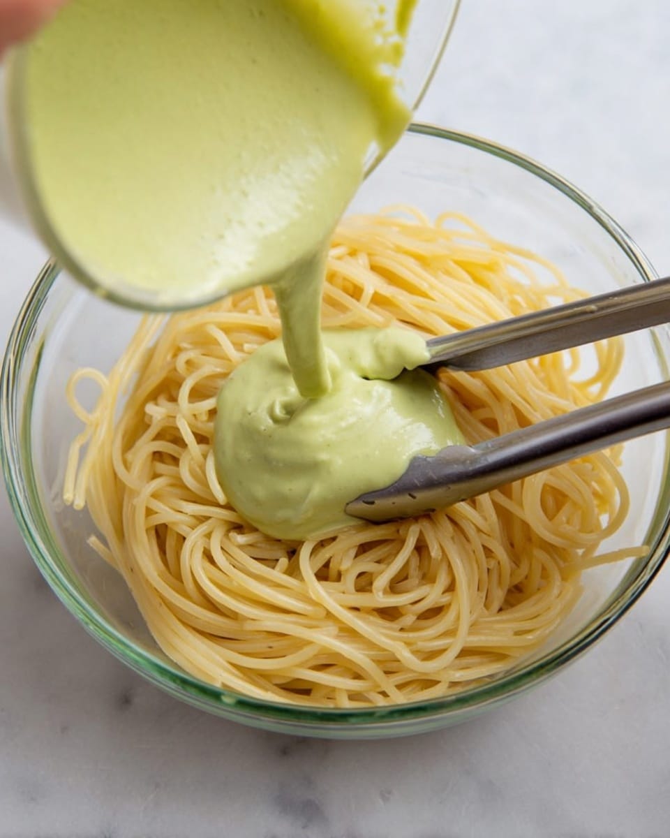 A clear glass bowl filled with light yellow spaghetti noodles sits on a white marbled surface. A woman's hand is pouring a thick, pale green sauce over the spaghetti from a blender or jug, creating a smooth layer of sauce on top of the noodles. Silver tongs rest inside the bowl among the noodles, partly lifted as if ready to mix the dish. The image shows a close, top-down view with soft lighting. photo taken with an iphone --ar 4:5 --v 7