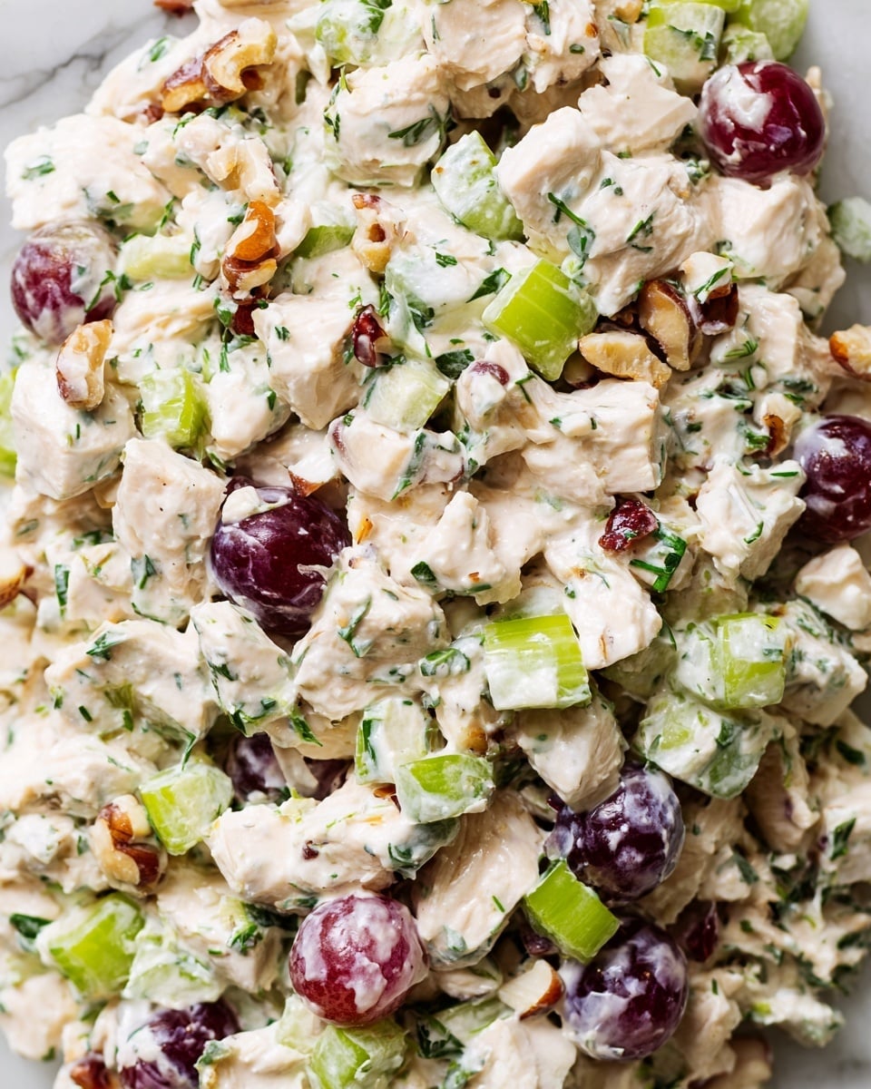 A close-up view of a creamy chicken salad mixture with visible chunks of white chicken meat combined with small pieces of bright green celery, halved purple-red grapes, and some toasted nuts. The mixture is coated in a thick, white dressing with small bits of green herbs scattered throughout. The salad appears fresh and textured with the different shapes and colors evenly spread. The whole dish is set against a white marbled surface. photo taken with an iphone --ar 4:5 --v 7