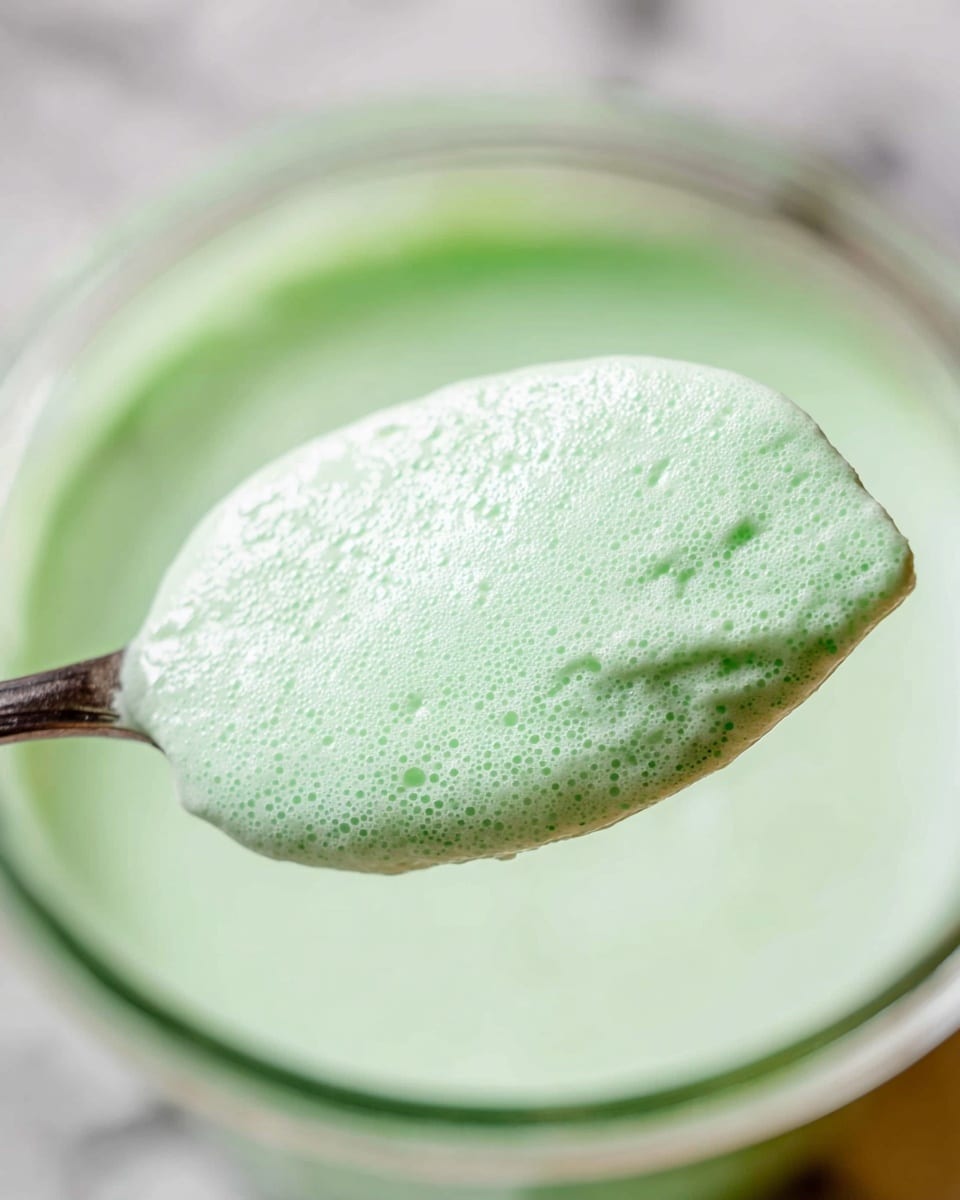 A close-up of a silver spoon holding a thick, foamy light green mixture with a smooth but slightly bubbly texture. The spoon is above a clear container filled with the same pale green mixture. The background is softly blurred with a white marbled texture visible below the container. The lighting highlights the creamy texture and the light reflections on the spoon and mixture. photo taken with an iphone --ar 4:5 --v 7