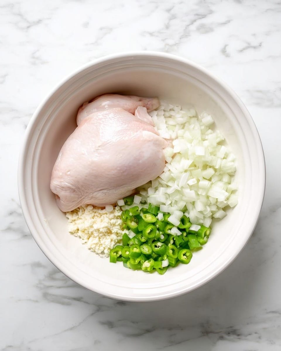 In a white bowl placed on a white marbled surface, there is one large raw chicken piece centered slightly left, surrounded by chopped white onions covering the right side of the bowl and some chopped green peppers positioned below the chicken piece. To the left of the green peppers, there is a small pile of minced garlic. The background and the bowl provide a clean, simple setting that highlights the colors and textures of the fresh ingredients. photo taken with an iphone --ar 4:5 --v 7
