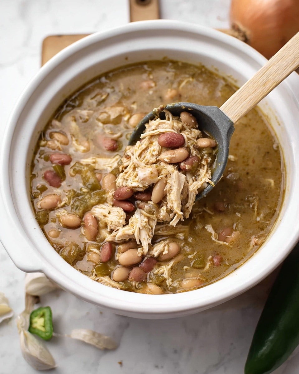 A white bowl filled with a thick soup made of light brown broth, shredded light tan chicken pieces, and various beans in shades of beige and reddish-brown. Small bits of green vegetables are scattered throughout, adding color contrast. A light wooden spoon with a dark gray scoop is lifting a spoonful of the soup, showing the chunky texture of the chicken and beans inside. The bowl sits on a white marbled surface, and parts of an onion, a jalapeño, and garlic are placed nearby around the bowl. Photo taken with an iphone --ar 4:5 --v 7