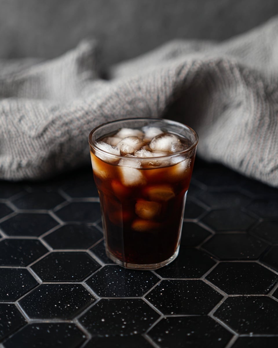 A clear glass filled with dark brown iced coffee, filled with many round and partly melted ice cubes that create a mix of white and brown areas inside the glass, is placed on a black hexagonal tile surface with subtle white specks. In the background, there is a soft, light gray knitted cloth with a slightly wrinkled texture. The overall setting has a calm, cozy feel with soft lighting that creates gentle reflections on the glass. photo taken with an iphone --ar 4:5 --v 7
