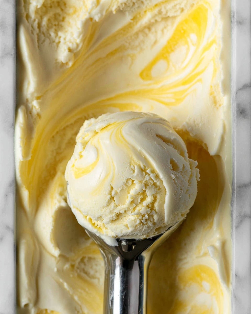The image shows a close-up of a single scoop of light yellow and cream ice cream held by a metal ice cream scoop. The ice cream has a smooth and creamy texture with visible swirls of yellow throughout the pale cream base. The scoop is centered in the image above a container filled with the same ice cream, which has a swirled and slightly uneven surface, showing both the creamy pale color and the brighter yellow streaks. The background is a white marbled texture. photo taken with an iphone --ar 4:5 --v 7