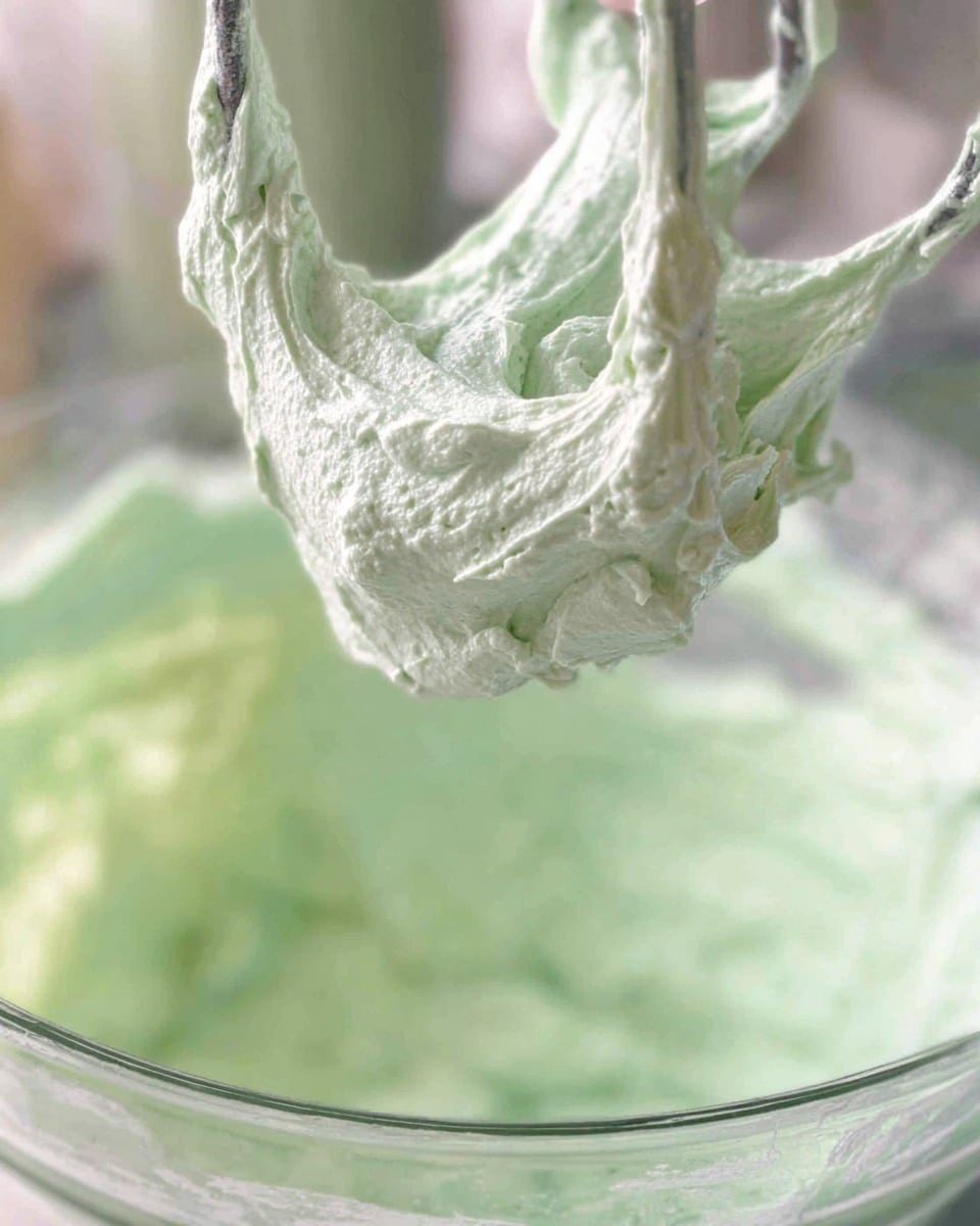 The image shows a close-up of thick, fluffy light green cream or frosting being lifted by a mixer attachment from a clear glass bowl. The texture looks soft and smooth with some peaks and swirls. The cream fills the bowl halfway, and the background is softly blurred. The overall color is pale green with a slightly whipped, airy look. Photo taken with an iphone --ar 4:5 --v 7