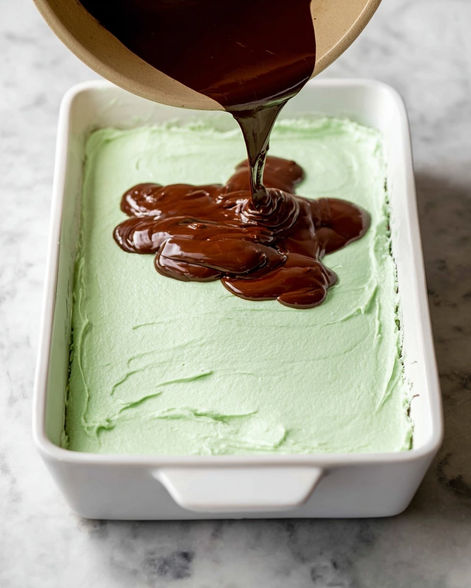 A white rectangular baking dish has two visible layers of dessert; the bottom layer is covered with a smooth, light green frosting that has some soft texture and spread marks, and on top of that, a thick, shiny dark brown chocolate sauce is being poured from a light-colored bowl into the center of the dish, creating a thick splash shape on the green layer. The dish sits on a white marbled surface. Photo taken with an iphone --ar 4:5 --v 7
