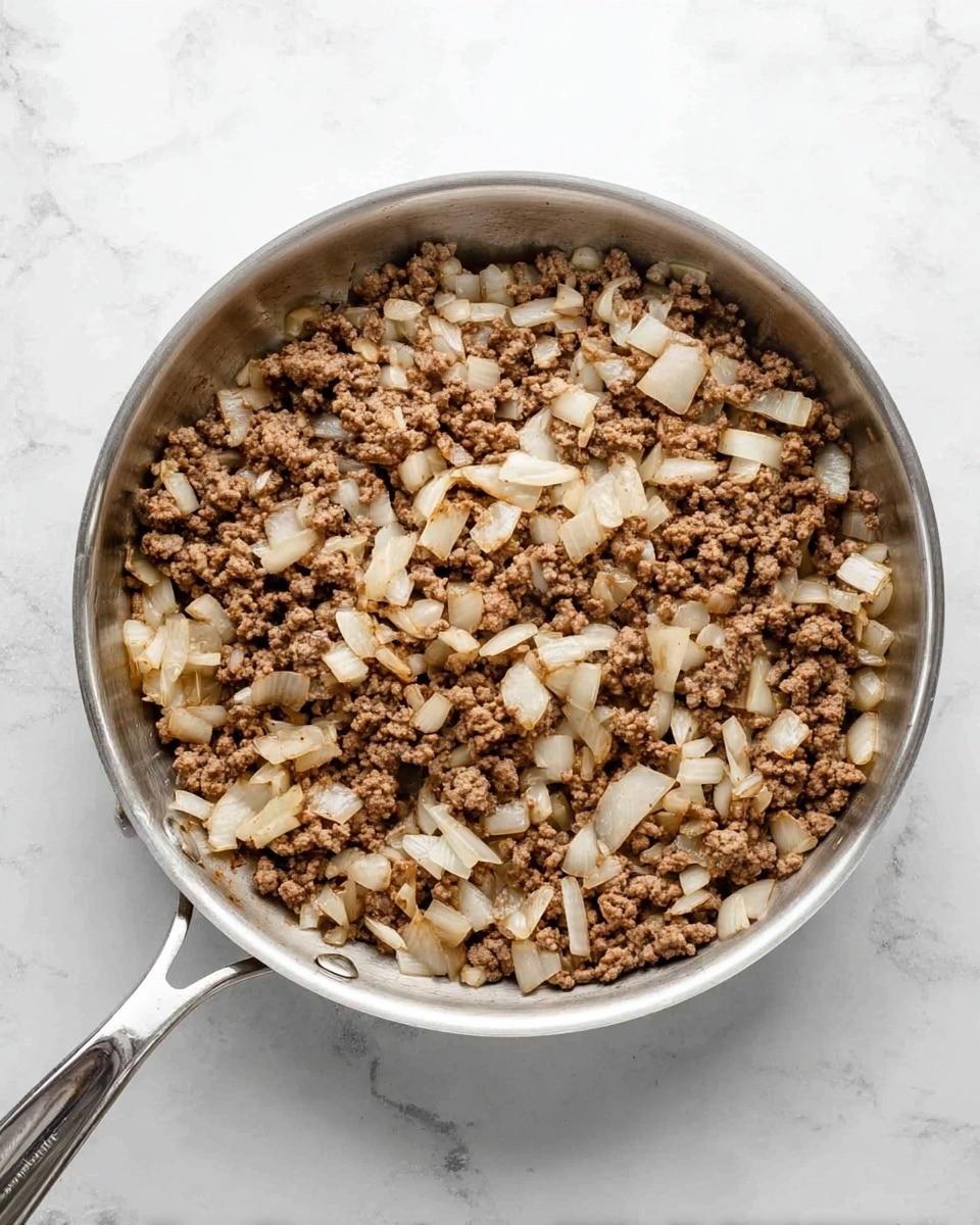 A top view of a silver frying pan filled with cooked ground meat and diced white onions, both mixed together. The meat looks brown and crumbly, while the onions are soft and translucent. The pan has a silver handle and is placed on a white marbled surface. photo taken with an iphone --ar 4:5 --v 7
