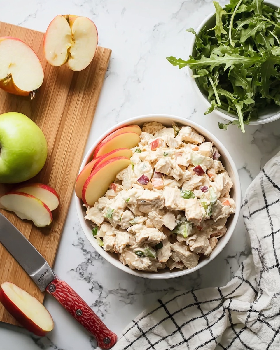A small white bowl filled with a soft textured, creamy salad that is light beige color with visible pieces of red and green bits mixed in. The salad sits on a bed of fresh, dark green leafy greens that fill the bowl's base and edges. On one side, there are five thin slices of vibrant red and yellow apple, neatly arranged in a row leaning on the salad. The bowl is placed on a white marbled surface, seen from above. Photo taken with an iphone --ar 4:5 --v 7