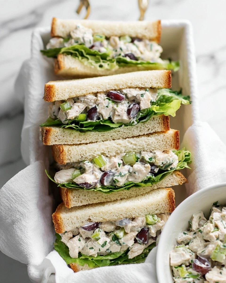 The image shows four sandwich halves neatly placed on a soft white cloth inside a white tray on a white marbled surface. Each sandwich has three layers: the top and bottom are light golden toasted bread slices with a soft texture, and the middle layer is a creamy chicken salad mixed with green leafy lettuce, small green herbs, and slices of dark purple grapes. The chicken salad looks chunky with visible pieces of white chicken and some green onion bits scattered throughout, adding texture and color contrast. On the side, there is a white bowl filled with extra creamy chicken salad containing similar ingredients. The overall look is fresh and inviting. photo taken with an iphone --ar 4:5 --v 7