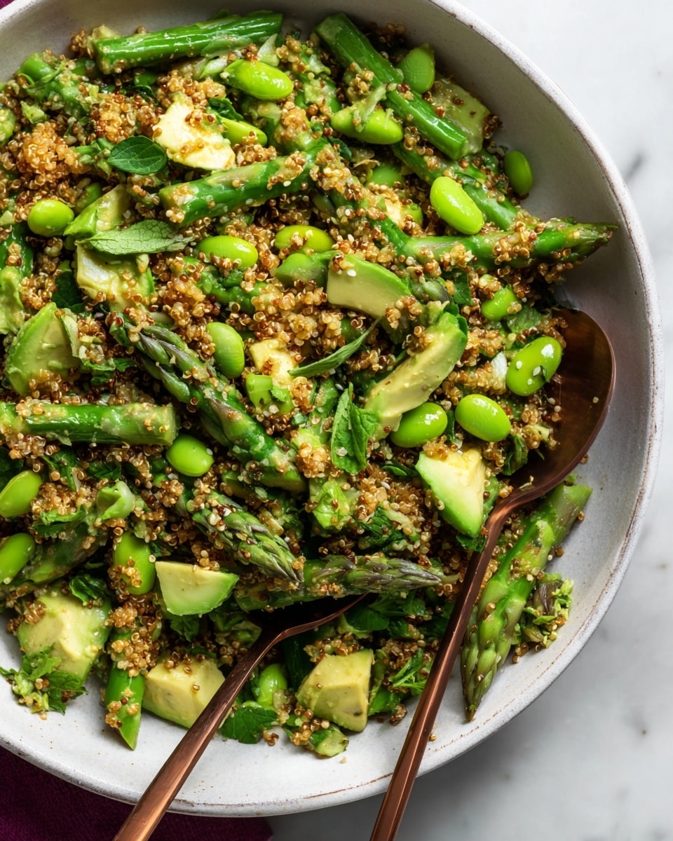 A close-up view of a salad in a large white bowl, showing three main layers: the base is bright green asparagus spears cut into pieces, with smooth-edged, light green avocado chunks scattered throughout. On top, medium brown quinoa grains are mixed evenly, giving a textured and grainy look, sprinkled with small sesame seeds. Some dark green leafy herbs and edamame beans add more color contrast and fullness. Two bronze spoons rest inside the bowl, partially submerged in the salad. The background is a white marbled surface with soft natural light highlighting the fresh colors. Photo taken with an iphone --ar 4:5 --v 7