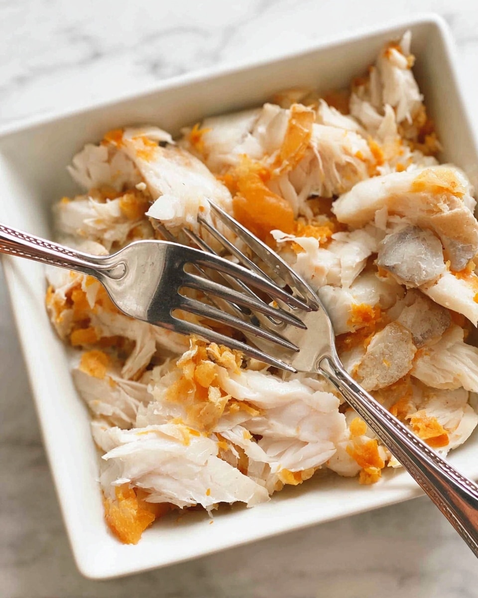 A white square dish is filled with torn pieces of cooked white fish that have a flaky texture and some patches of bright orange skin still attached. Two silver forks are poking into the fish, showing a mix of smooth white flesh and rough orange skin scattered evenly across the dish. The background is a white marbled surface. Photo taken with an iphone --ar 4:5 --v 7