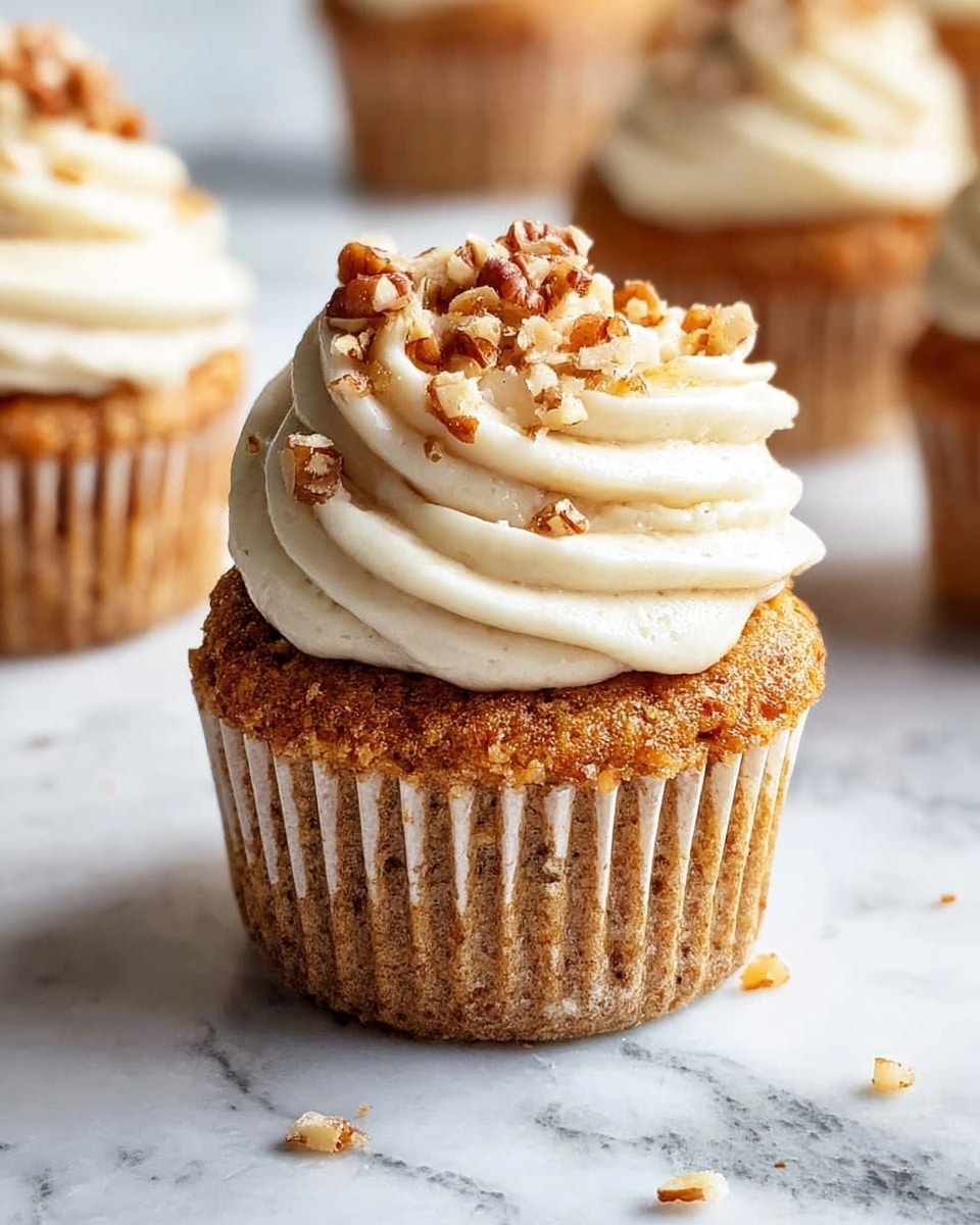 A close-up view of a cupcake with two visible layers: the bottom layer is light brown and textured with small nut pieces, wrapped in a light beige paper liner; the top layer is a thick swirl of creamy white frosting crowned with small chopped nuts scattered on top. The cupcake is placed on a white marbled surface with a few nut bits around it, and blurred cupcakes are seen in the background. photo taken with an iphone --ar 4:5 --v 7