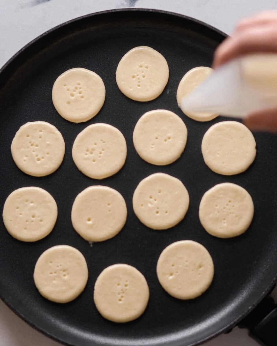 A close-up image shows many small, round dollops of smooth, light beige batter evenly spaced on a black skillet surface. The batter forms perfect circles about the same size, with a few tiny bubbles appearing on the tops, indicating cooking in progress. On the right side, a woman's hand holds a clear piping bag, gently squeezing to add another small batter circle. The background is a white marbled texture. photo taken with an iphone --ar 4:5 --v 7