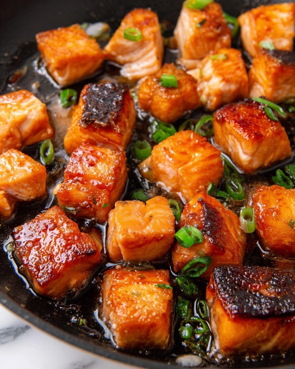 The image shows many pieces of cooked salmon in a pan, each piece cut into small cubes with a golden brown and slightly crispy outside layer. Some pieces have the darker salmon skin on one side. The salmon chunks have a shiny, glazed look with a caramelized texture, and small green slices of chopped green onion are sprinkled on top and between the pieces. The background is a black pan with a little oil reflecting light, all on a white marbled surface. photo taken with an iphone --ar 4:5 --v 7