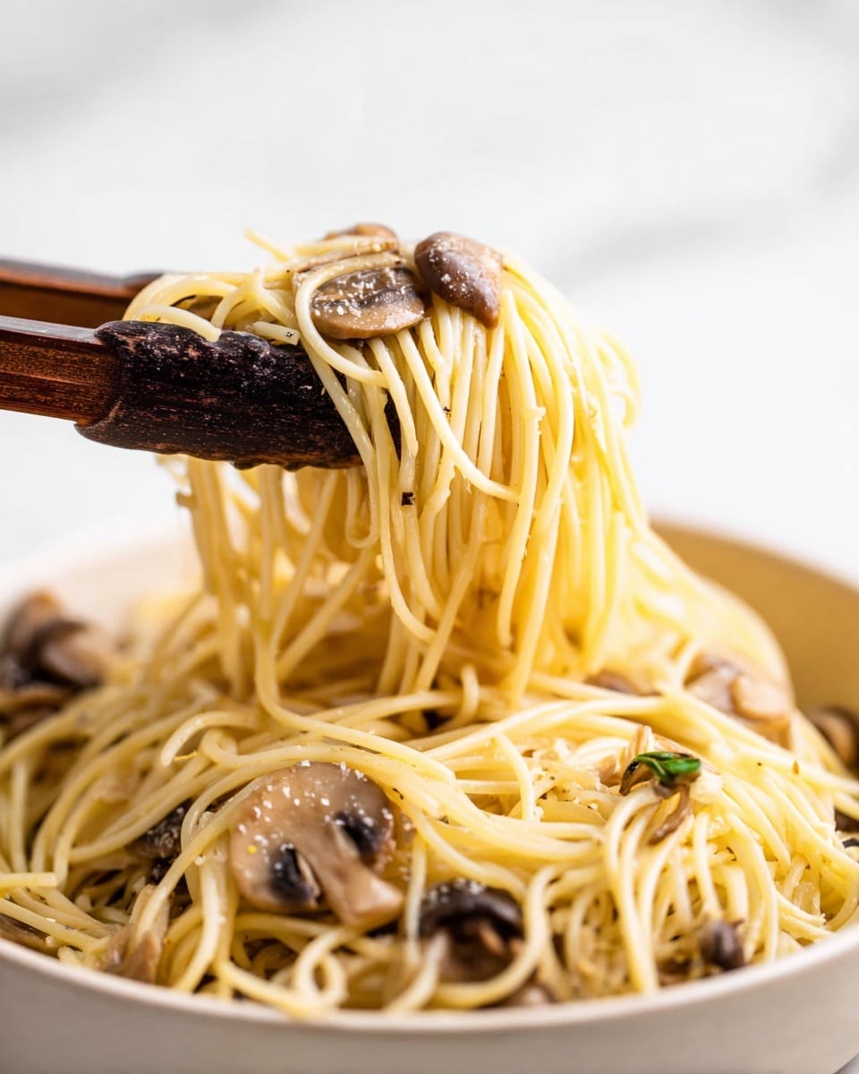 A close-up of long light yellow spaghetti noodles mixed with small brown mushroom slices sits in a white bowl. A pair of dark wooden tongs lifts a twisted bundle of noodles with mushrooms above the bowl. The spaghetti appears soft and slightly glossy, resting on a white marbled surface. The background is bright white, emphasizing the noodles and mushrooms. photo taken with an iphone --ar 4:5 --v 7