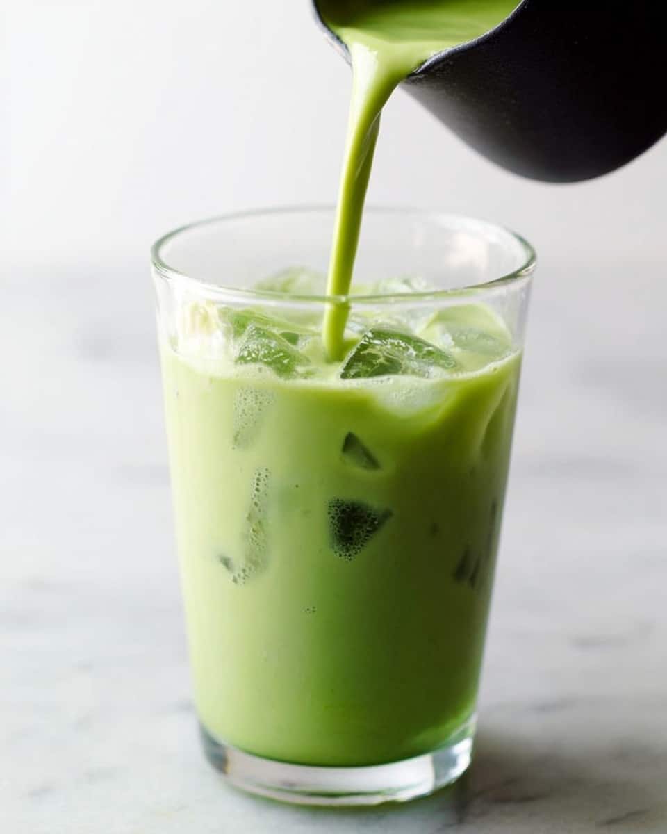 A clear glass filled almost to the top with light green iced matcha latte displaying a smooth creamy texture and visible ice cubes floating inside. A small amount of frothy bubbles gathers on the surface. A woman's hand is pouring a fresh stream of vivid green matcha liquid from a black container into the glass. The background and surface are white marble, giving a clean and bright look. photo taken with an iphone --ar 4:5 --v 7
