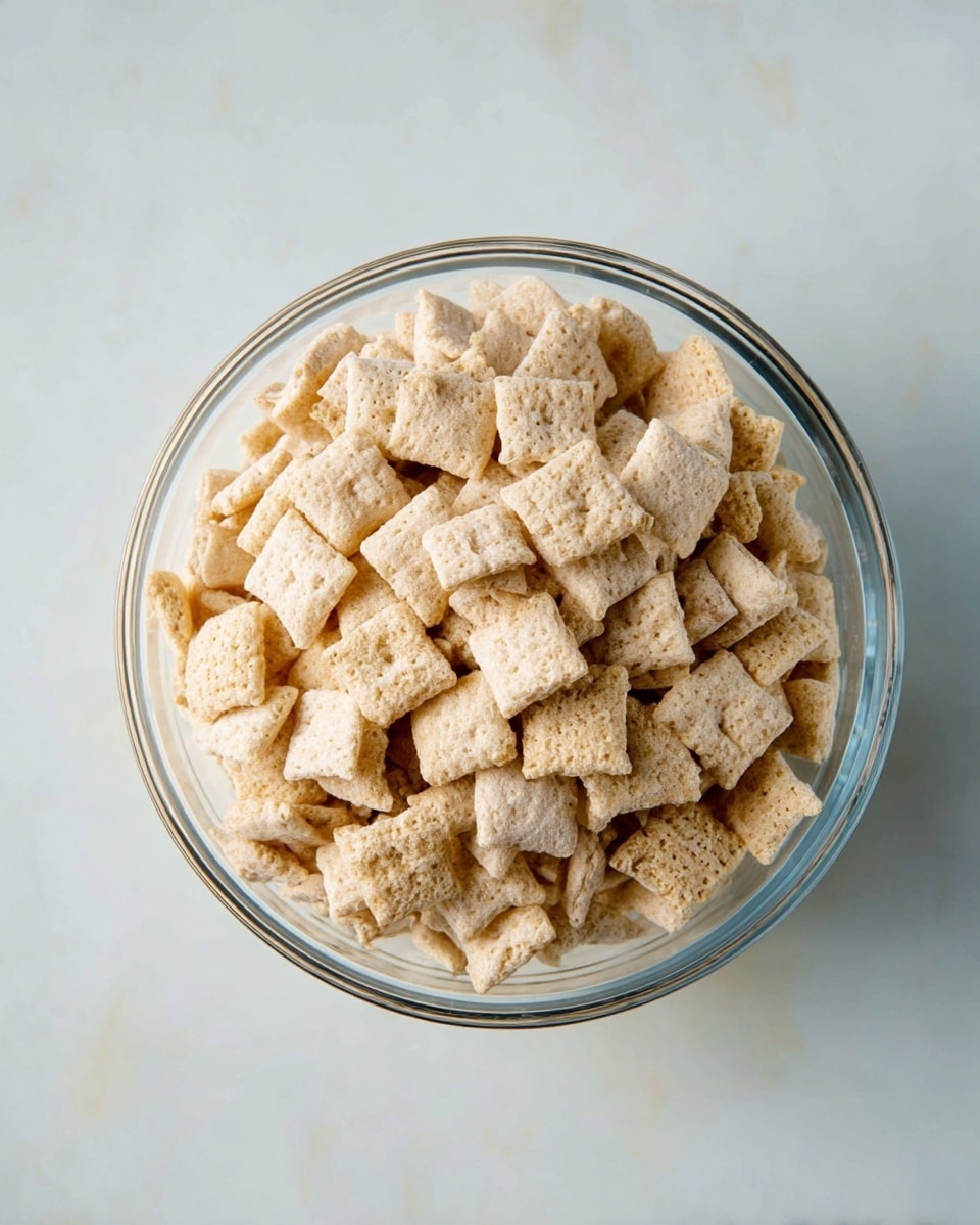 A clear glass bowl filled with light beige square cereal pieces, each piece showing a slightly rough texture with small holes and some variation in shape. The bowl sits on a white marbled surface with soft lighting that highlights the cereal's dry, crunchy look. The cereal pieces are heaped to the top of the bowl, creating a thick single layer that almost spills over the edge. photo taken with an iphone --ar 4:5 --v 7