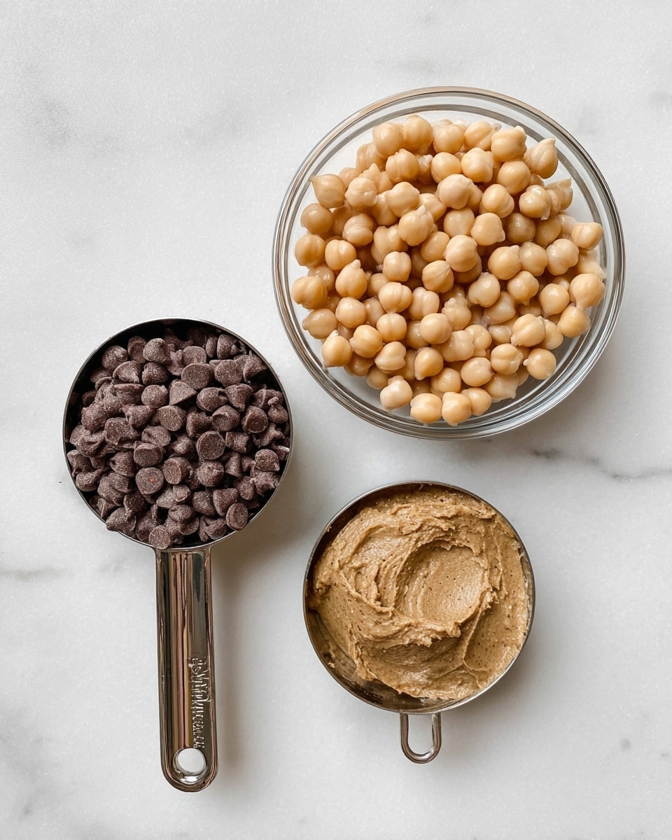 The image shows three containers of ingredients placed on a white marbled textured surface. On the top right, there is a clear glass bowl filled with round, beige chickpeas. On the bottom left, there is a shiny metal measuring cup, filled to the brim with dark brown chocolate chips. On the bottom right, another shiny metal measuring cup contains a smooth light brown nut butter with a slightly grainy texture. Photo taken with an iphone --ar 4:5 --v 7