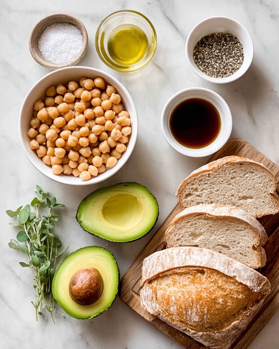 The image shows a white bowl full of round light brown chickpeas placed near the center, with two halves of a green avocado, one half showing a large brown seed, positioned below it. To the top right of the chickpeas, there are two small white bowls, one with a dark brown liquid and the other with finely ground black pepper. Below these, another small white bowl has coarse white salt. To the left of the chickpeas, there are a few fresh green oregano sprigs. At the bottom right of the image, there is a wooden board holding a loaf of sliced bread with a golden brown crust and a porous light beige inside. Everything is placed on a white marbled textured surface. Photo taken with an iphone --ar 4:5 --v 7